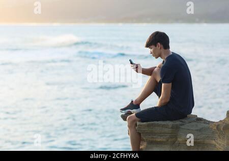 Jeune garçon utilisant un téléphone portable assis sur le bord d'une falaise avec vue sur l'océan en arrière-plan pendant le coucher du soleil. Communication et jeunesse. Banque D'Images