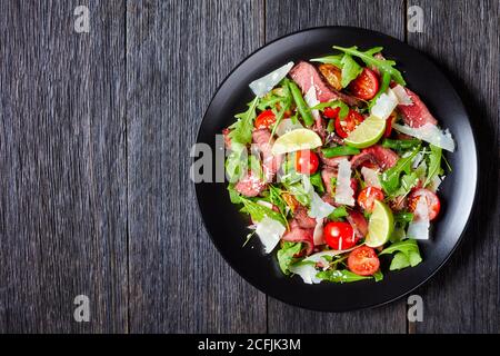 Salade de tagliata au bœuf en tranches avec haricots verts, tomates cerises, arugula fraîche et quartiers de parmesan et de citron vert, servi sur une assiette noire sur un bac en bois rustique Banque D'Images