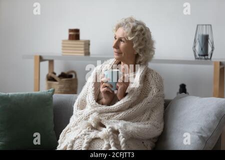 Une femme mûre et mûre rêveuse a enveloppé une couverture chaude pour se détendre sur le canapé Banque D'Images