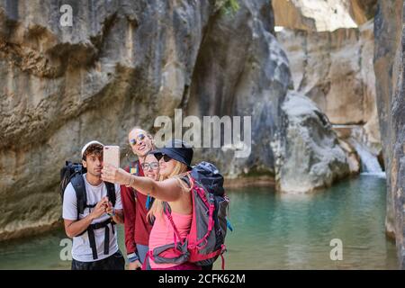 Groupe d'amis enchantés avec des sacs à dos emportant le selfie sur un smartphone ensemble en se tenant dans le canyon près du lac pendant les vacances d'été Banque D'Images