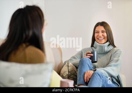 Des femmes ravies avec des tasses de café assis sur le canapé salon et rire à plaisanter ensemble tout en divertissant pendant week-end Banque D'Images