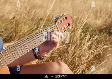 Crop jeune homme guitariste talentueux assis dans un pré séché et jouer d'un instrument de musique acoustique par beau temps Banque D'Images