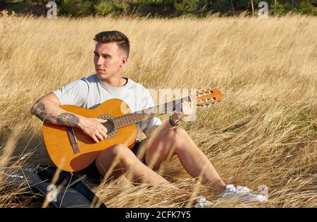 Jeune guitariste masculin talentueux assis dans un pré séché et jouant instrument de musique acoustique le jour ensoleillé Banque D'Images