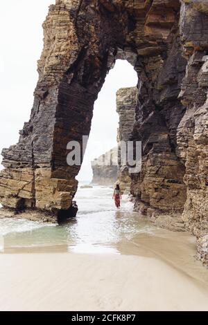 Vue lointaine de la femme en voyage debout dans l'eau de mer sous formation de pierre en forme d'arche Banque D'Images