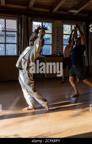 Vue latérale du corps entier d'un instructeur masculin méconnaissable qui fait du chi mouvement de kung ou qigong avec les bras levés pendant la pratique avec étudiants Banque D'Images