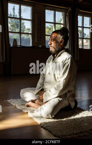 Vue latérale complète du corps d'un homme senior ethnique barbu pratiquer la méditation chi kung en position lotus dans un studio sombre Banque D'Images