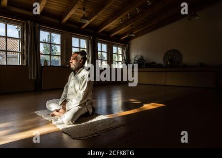 Vue latérale complète du corps d'un homme senior ethnique barbu pratiquer la méditation chi kung en position lotus dans un studio sombre Banque D'Images