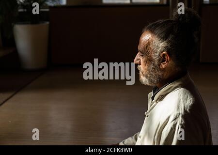 Récolte de l'homme senior ethnique barbu focalisé pratiquant le chi kung méditation en position lotus dans un studio sombre Banque D'Images