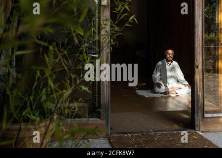 Vue de face du corps entier d'un homme senior ethnique barbu pratiquer la méditation chi kung en position lotus dans un studio sombre Banque D'Images