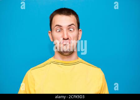 Drôle homme de bande dessinée fait grimace avec des croix yeux. Jeune homme en t-shirt jaune avec l'expression folle a l'amusement seul, joue imbécile isolé sur le dos bleu Banque D'Images