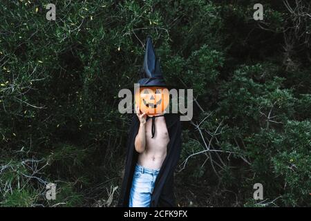 Enfant méconnaissable dans un manteau noir d'Halloween couvrant le visage avec de l'air ballon en forme de citrouille Banque D'Images