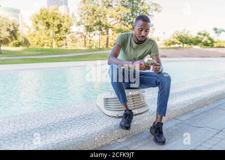 Homme afro-américain pensif assis à la frontière près de la fontaine dedans La ville et surfer sur Internet sur smartphone tout en vous refroidissant pendant Banque D'Images