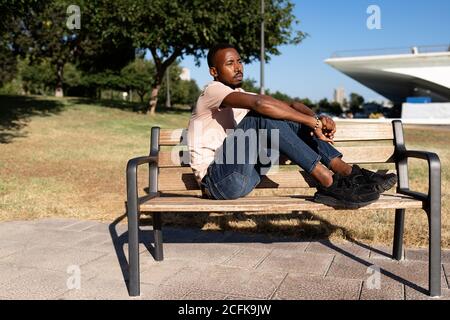 Vue latérale d'un homme afro-américain décontracté assis sur le banc dans un parc urbain et à genoux tout en regardant loin Banque D'Images