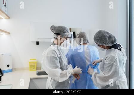 Les femmes travailleurs médicaux dans les masques de protection et les écrans faciaux aider les uns les autres pour s'habiller pendant la préparation à l'intervention médicale à l'hôpital Banque D'Images