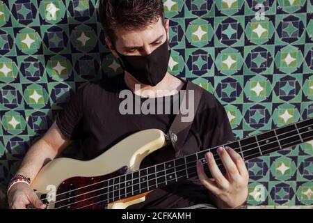 Corps complet du jeune homme en masque de protection noir pour prévention des coronavirus assis sur des marches près du mur et jouant de la basse guitare dans la rue Banque D'Images