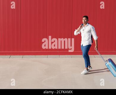 Corps complet de jeune homme afro-américain dans élégant tenue portant des bagages tout en marchant contre le mur rouge dans la rue Banque D'Images