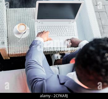 crop anonyme homme d'affaires noir en costume formel assis à table avec une tasse de café et la saisie sur un ordinateur portable tout en travaillant dans le café Banque D'Images