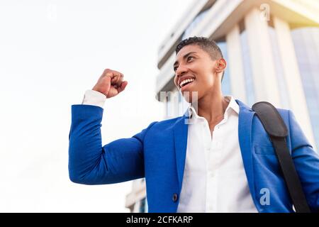 Faible angle de jeune homme afro-américain joyeux dans costume bleu formel célébrant le succès tout en se tenant contre le moderne bâtiment de la ville Banque D'Images