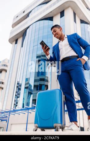 De dessous de positif jeune homme d'entrepreneur afro-américain dans combinaison élégante à proximité d'une valise et utilisation de l'application pour appeler taxi dans une rue moderne Banque D'Images