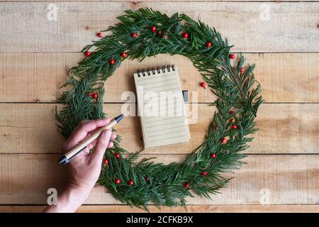 Vue de dessus de Crop anonyme femme avec stylo écriture menu de fête dans le carnet placé à l'intérieur de la couronne de Noël sur la table en bois Banque D'Images