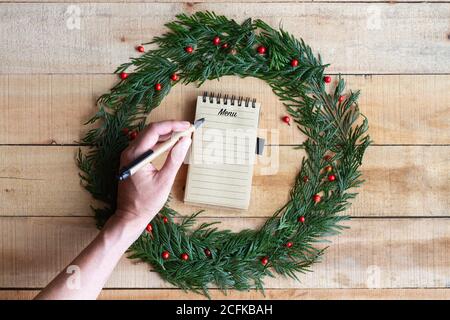 Vue de dessus de Crop anonyme femme avec stylo écriture menu de fête dans le carnet placé à l'intérieur de la couronne de Noël sur la table en bois Banque D'Images