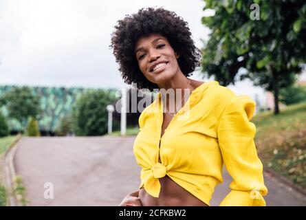 Femme noire optimiste dans des vêtements branchés souriant pour la caméra et dansant avec les bras levés le jour ensoleillé dans le parc Banque D'Images