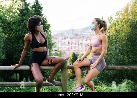 Jeunes filles en forme d'oreilles actives et masques de protection assis sur une clôture en bois et bavarder en faisant une pause en plein air formation en été Banque D'Images