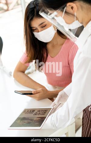 Un client asiatique scanne le menu en ligne du code QR depuis une serveuse avec masque et masque facial. Le client s'est assis sur une table de distanciation sociale pour un nouveau style de vie normal Banque D'Images