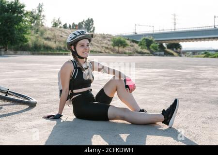 Vue latérale en grand angle de la jeune femme heureuse en vêtements de sport et casque avec sac à dos assis sur un sol pavé et reposant après avoir fait du vélo en été Banque D'Images