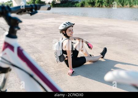 Vue latérale en grand angle de la jeune femme heureuse en vêtements de sport et casque avec sac à dos assis sur un sol pavé et reposant après avoir fait du vélo en été Banque D'Images