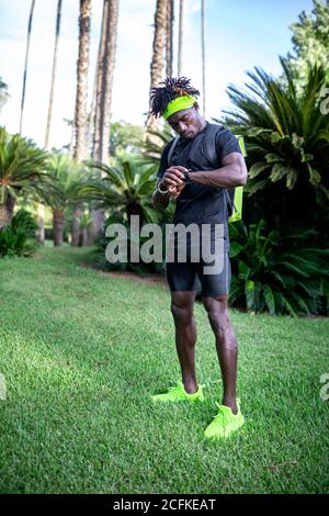 Jeune coureur afro-américain élégant avec casque sur le cou vérification des résultats sportifs sur le tracker d'entraînement en vert parc tropical Banque D'Images