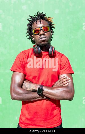 Athlète afro-américain fatigué dans les vêtements de sport avec casque relaxant dans la rue après l'entraînement le jour ensoleillé regardant la caméra Banque D'Images