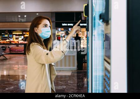 une femme avec un masque utilise le distributeur automatique pour acheter une collation Banque D'Images