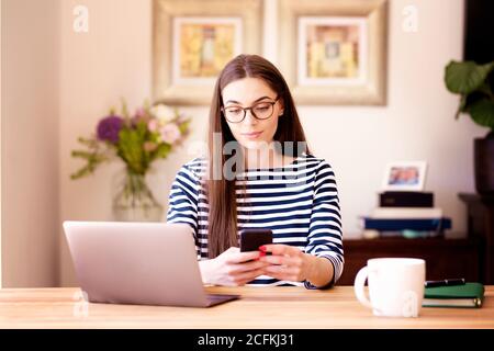 Portrait-it tourné de la messagerie texte de jeune femme souriante tout en étant assis au bureau et en travaillant de la maison. Bureau à domicile. Banque D'Images