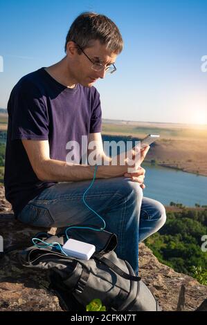 Man On a rando utilise un smartphone tout en se chargeant depuis la banque d'énergie sur le rocher à l'aube. Banque D'Images