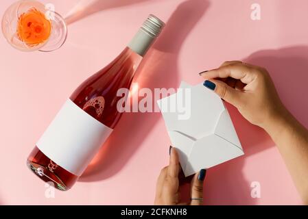 Carte postale de voeux avec bouteille de vin rose et carte d'enveloppe dans les mains de la femme sur un fond rose clair avec des ombres, espace de copie. Vue de dessus. Banque D'Images