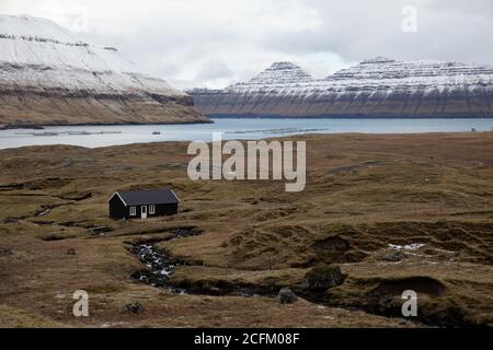 Paysage à couper le souffle de maison résidentielle en bois située près de la rivière dans vallée des highlands avec des montagnes couvertes de neige sur les îles Féroé Banque D'Images