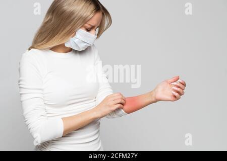Studio portrait de la jeune femme malade dans un masque protecteur rayant son bras, isolé sur fond gris. Symptômes d'allergie animale, démangeaisons de la peau Banque D'Images