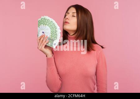 Satisfaite heureuse femme avec des cheveux bruns dans le chandail rose odeur ventilateur de centaines d'euros dans sa main, appréciant l'argent, la richesse. Prise de vue studio en intérieur isolée Banque D'Images