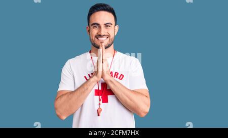 Jeune homme beau portant l'uniforme de maître-nageur priant avec les mains ensemble demandant pardon souriant confiant. Banque D'Images