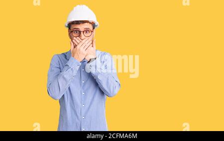 Beau jeune homme avec l'ours portant le casque d'architecte choqué couvrant la bouche avec les mains pour erreur. Concept secret. Banque D'Images