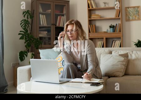 Femme d'affaires blonde mûre sérieuse en tenue décontractée assise sur le canapé petite table Banque D'Images