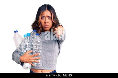 Jeune belle femme de course mixte tenant des bouteilles de plastique de recyclage pointant avec le doigt vers l'appareil photo et à vous, geste confiant regardant sérieux Banque D'Images