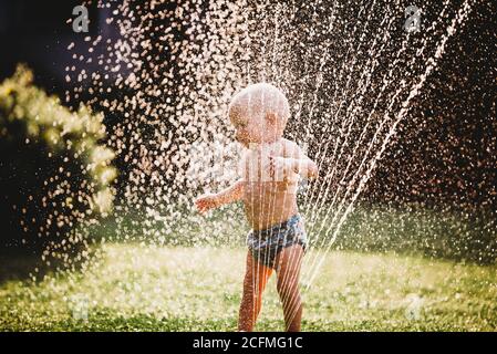 Bébé blanc debout dans le jardin jouant avec l'eau de la arroseur Banque D'Images