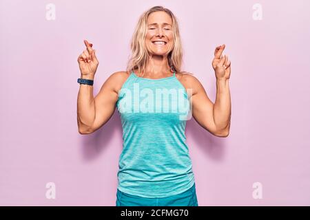 Femme blonde caucasienne d'âge moyen portant des vêtements de sport sur fond rose gesturant le doigt croisé souriant avec espoir et les yeux fermés. Chance et sup Banque D'Images