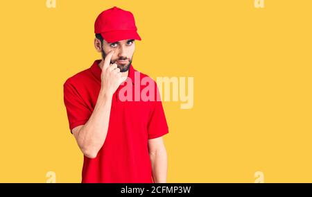 Jeune homme beau avec la barbe portant l'uniforme de livraison pointant vers l'œil vous regardant le geste, expression suspecte Banque D'Images