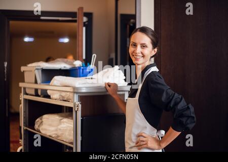 Une femme de ménage souriante avec un chariot de nettoyage et des fournitures de nettoyage, regardant la caméra et posant. Banque D'Images