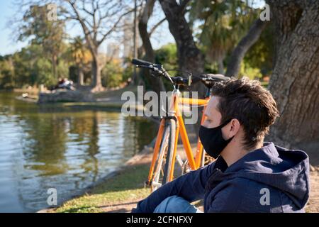 Jeune homme avec un masque en tissu reposant avec son vélo au bord d'un lac dans un parc à Buenos Aires, Argentine, un après-midi d'hiver ensoleillé, pendant l'épidémie de c Banque D'Images