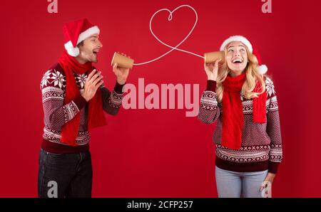 Message d'amour de Noël. Mignon couple en chapeau de père Noël et chandails parlant sur téléphone de boîte sur fond rouge Banque D'Images