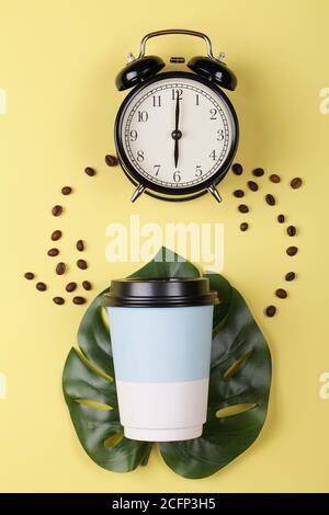 Tasse de café en papier sur une feuille de monstère et réveil sur fond jaune coloré. Concept de réveil du matin. Vue de dessus de la pose à plat Banque D'Images